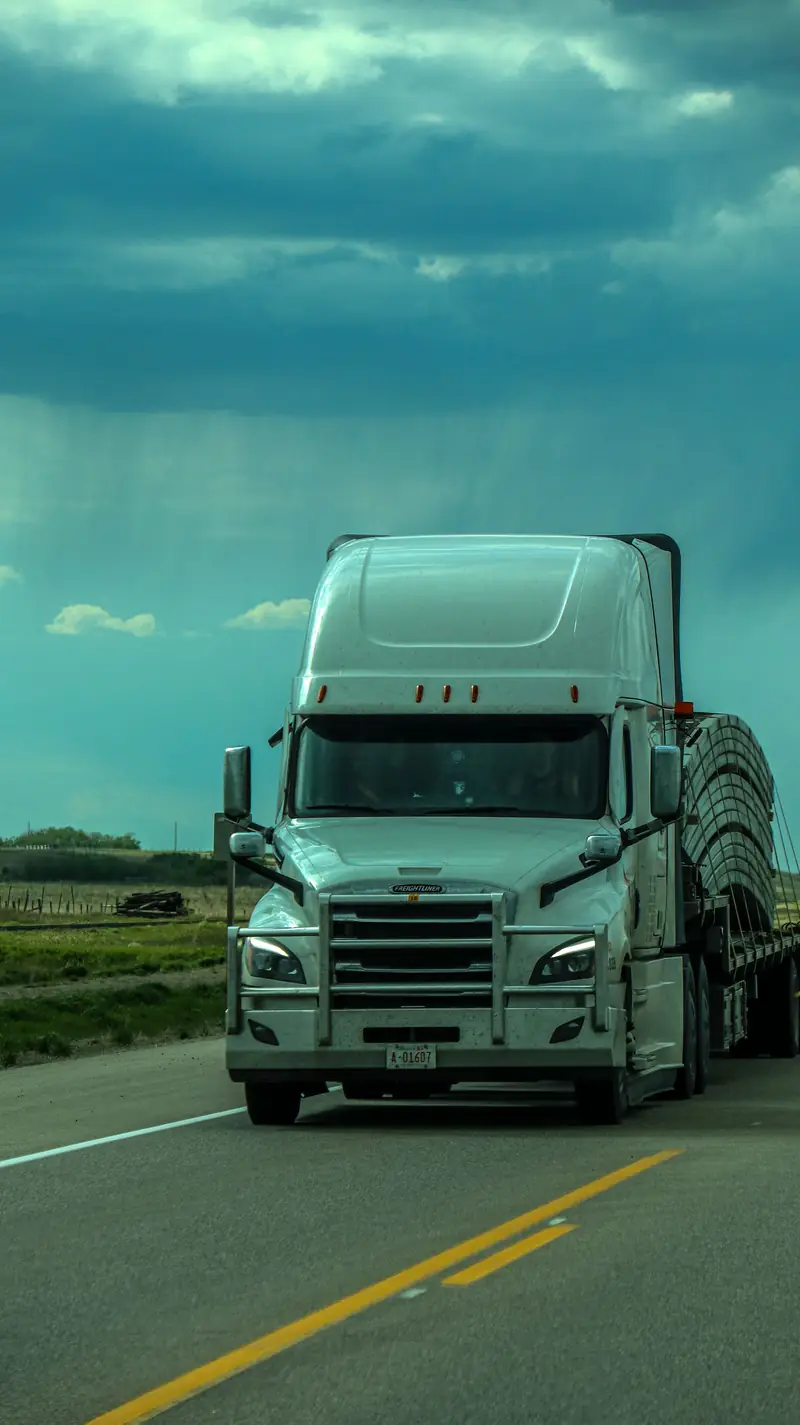 A white semi-truck hauling a covered flatbed load on a two-lane highway under a stormy sky